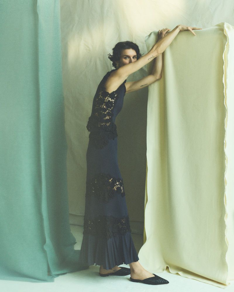 A woman poses in a sleeveless navy dress with lace details, leaning against a draped backdrop with muted tones in an indoor setting.