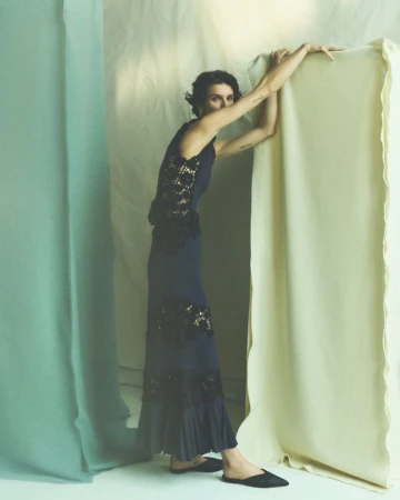 A woman poses in a sleeveless navy dress with lace details, leaning against a draped backdrop with muted tones in an indoor setting.