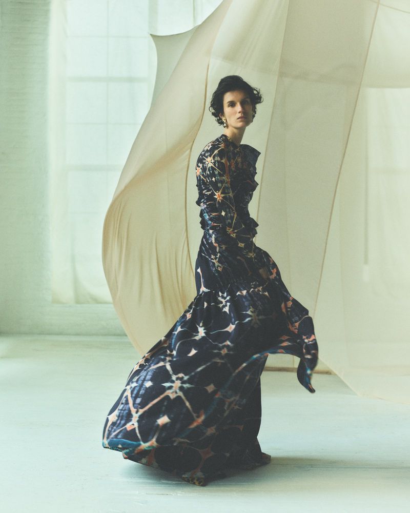Woman in patterned gown poses in studio near sheer draped fabric. Fashion shot with elegant dark and light dress and neutral background.