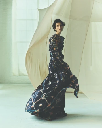 Woman in patterned gown poses in studio near sheer draped fabric. Fashion shot with elegant dark and light dress and neutral background.
