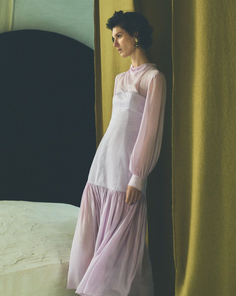 A woman wearing a light purple dress poses indoors in front of a dark wall with a yellow-gold fabric curtain, fashion portrait.