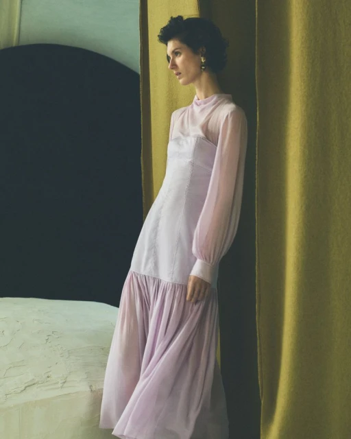 A woman wearing a light purple dress poses indoors in front of a dark wall with a yellow-gold fabric curtain, fashion portrait.