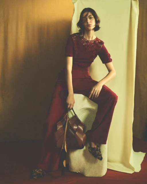 A woman in a red lace top and matching pants sits on a white block, holding a brown leather bag in a studio with warm lighting.