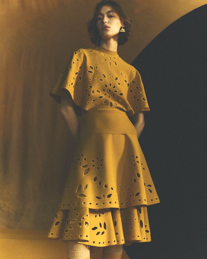 A young woman poses in a mustard-colored dress with floral laser-cut detailing, complemented by matching earrings and neutral stockings.