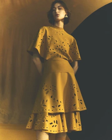 A young woman poses in a mustard-colored dress with floral laser-cut detailing, complemented by matching earrings and neutral stockings.