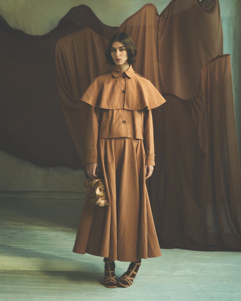 A woman models a brown skirt suit with capelet and gladiator sandals against a background of draped fabric in an indoor setting.