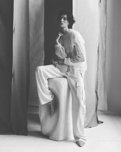 Elegant woman in sheer top and white cargo pants sits in studio surrounded by draped fabric in a black and white fashion portrait.