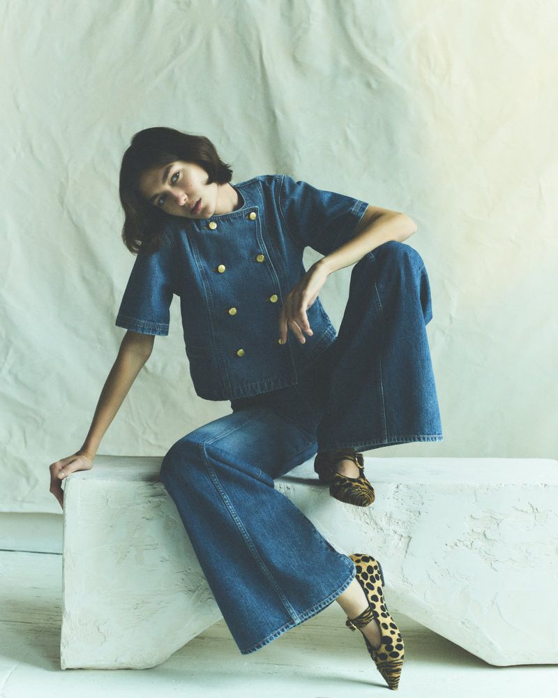 A stylish woman in a denim outfit and animal-print shoes sits in a neutral studio, striking a pose with a serious facial expression.