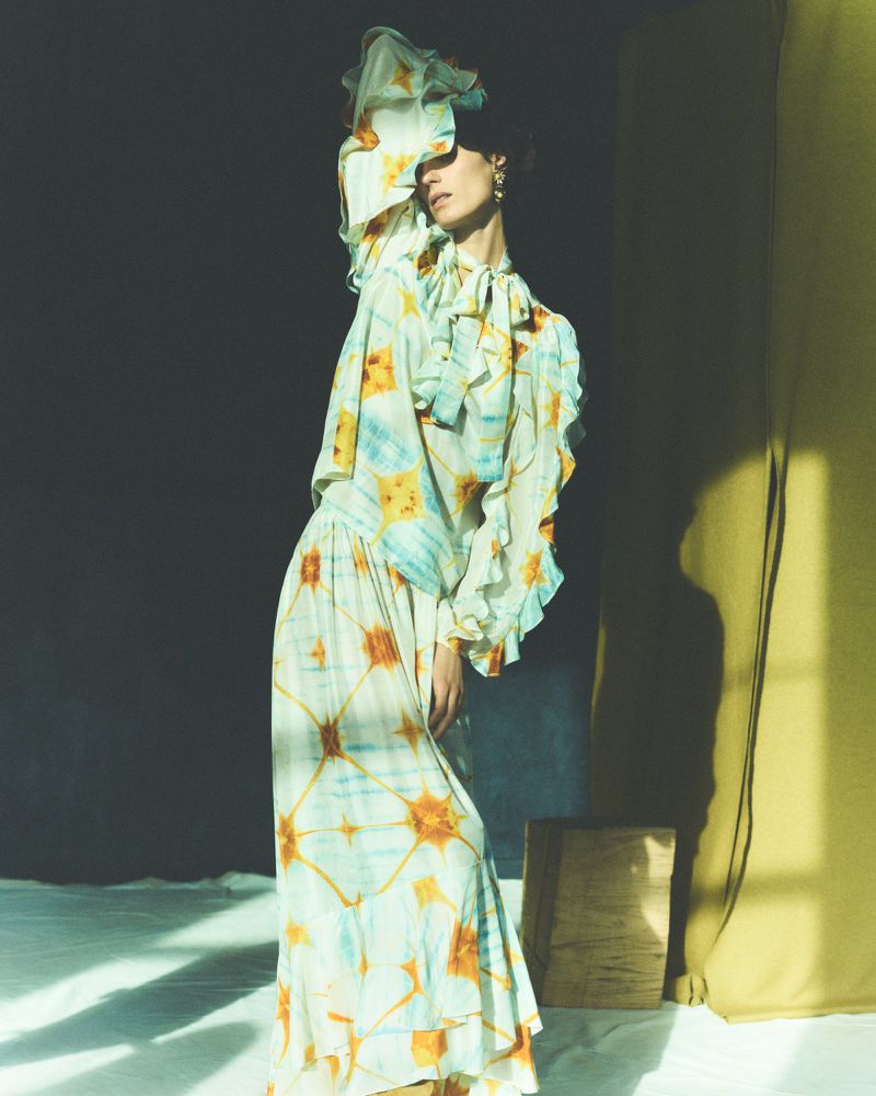 A woman in a floral dress poses in a photo studio with gray and yellow fabric backdrop, captured in soft, diffused light.