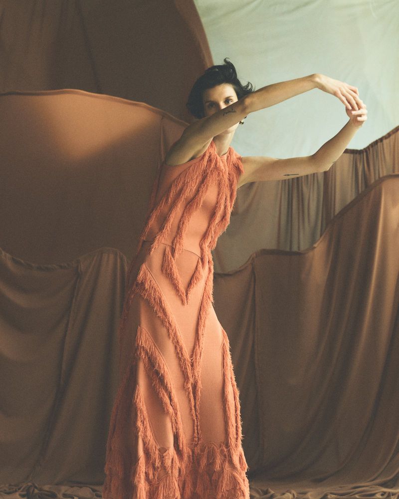 An adult woman in a flowing fringe dress stands in a dynamic pose, with arms raised against a backdrop of draped neutral-toned fabric.