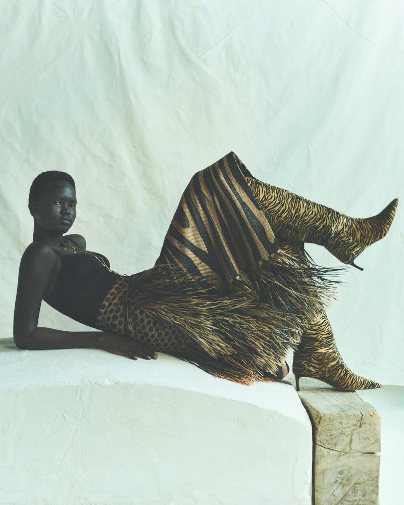 Striking portrait of an African woman in a black top, animal print skirt and knee high boots, lying on a white, rough platform indoors.