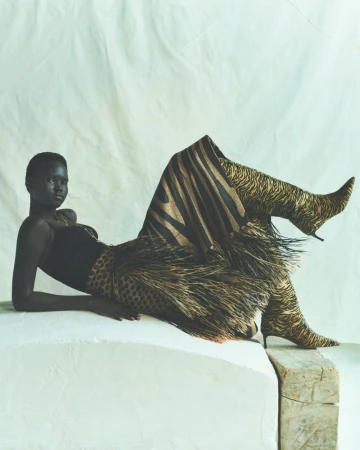 Striking portrait of an African woman in a black top, animal print skirt and knee high boots, lying on a white, rough platform indoors.