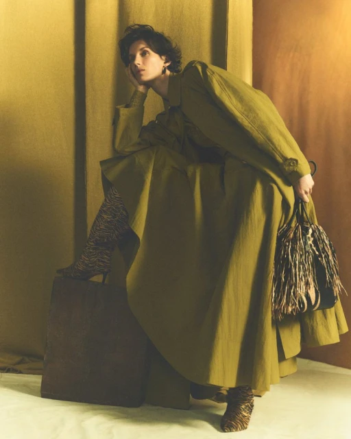 Olive-toned fashion shot of a woman in a coat and animal-print boots, seated on a block against a mottled brown and yellow backdrop.