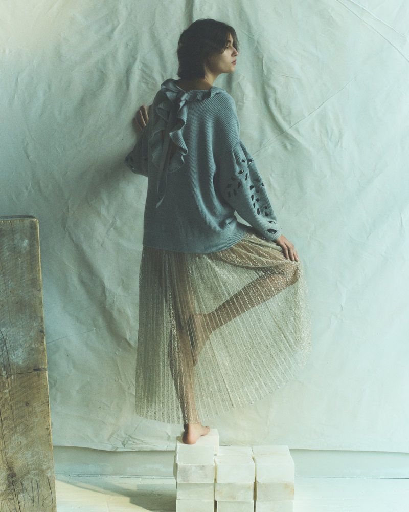 Woman in a grey sweater and gold pleated skirt posing on stone blocks against a draped cloth backdrop in a fashion editorial.