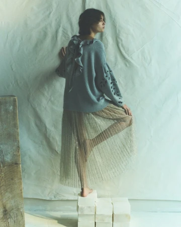 Woman in a grey sweater and gold pleated skirt posing on stone blocks against a draped cloth backdrop in a fashion editorial.