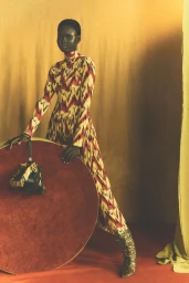 Model in patterned high-neck dress leaning on a large round prop against a warm draped backdrop