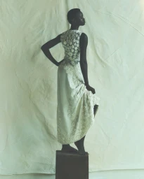 Model standing on a pedestal wearing a white lace dress against a crumpled cream backdrop.