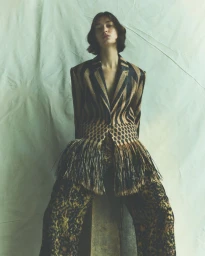 Model wearing a patterned striped jacket with fringe detail and dark patterned trousers against a textured background.