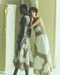 Two models wearing white and gold lace dresses posing together against a draped neutral fabric backdrop