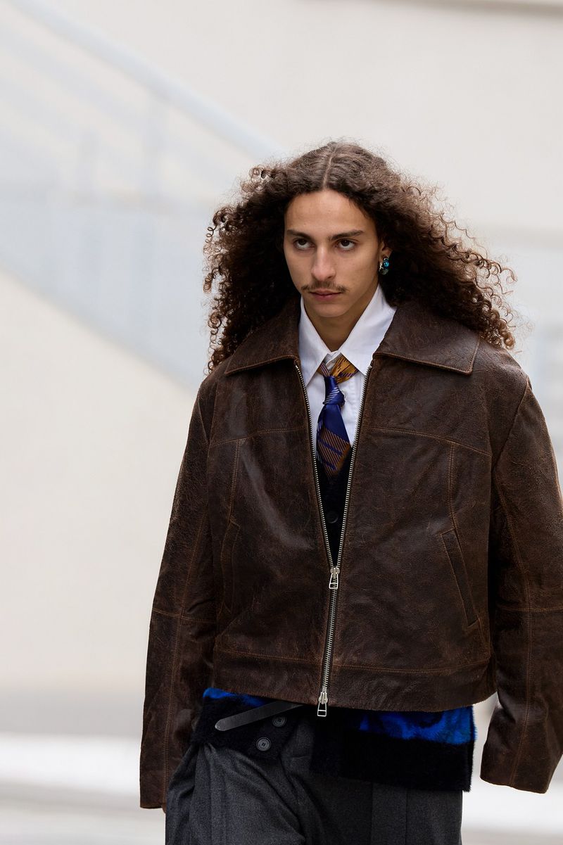 Runway fashion show: a single male model with curly hair wears a brown leather jacket, tie, grey pants, captured in medium shot.