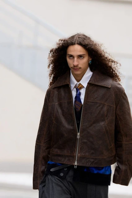Runway fashion show: a single male model with curly hair wears a brown leather jacket, tie, grey pants, captured in medium shot.