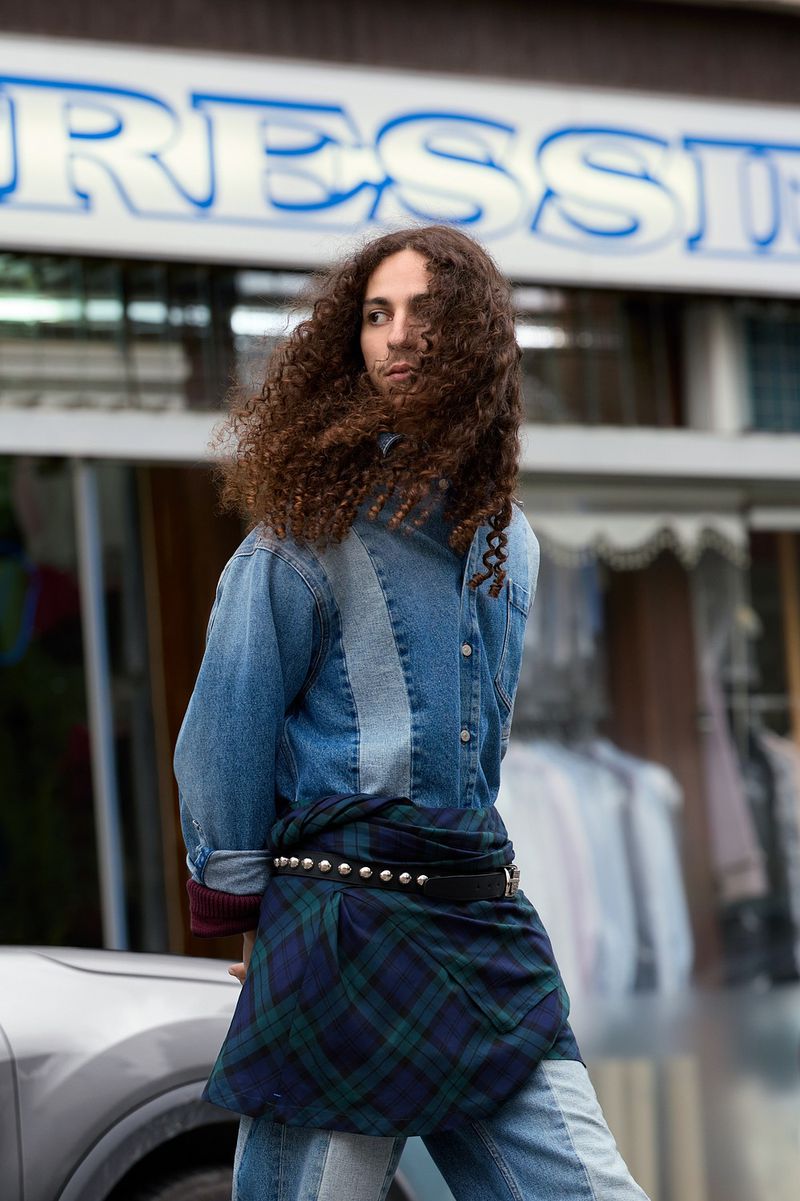 Adult male with curly brown hair stands in urban outdoor setting, wearing a denim jacket, kilt-like garment, and jeans, looking contemplative.