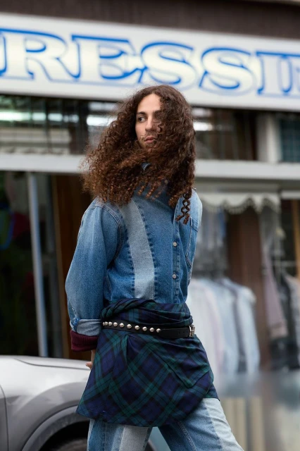 Adult male with curly brown hair stands in urban outdoor setting, wearing a denim jacket, kilt-like garment, and jeans, looking contemplative.