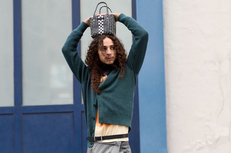 A young adult male with long curly hair balances a black-and-white woven bag on his head. He is wearing a green cardigan sweater.