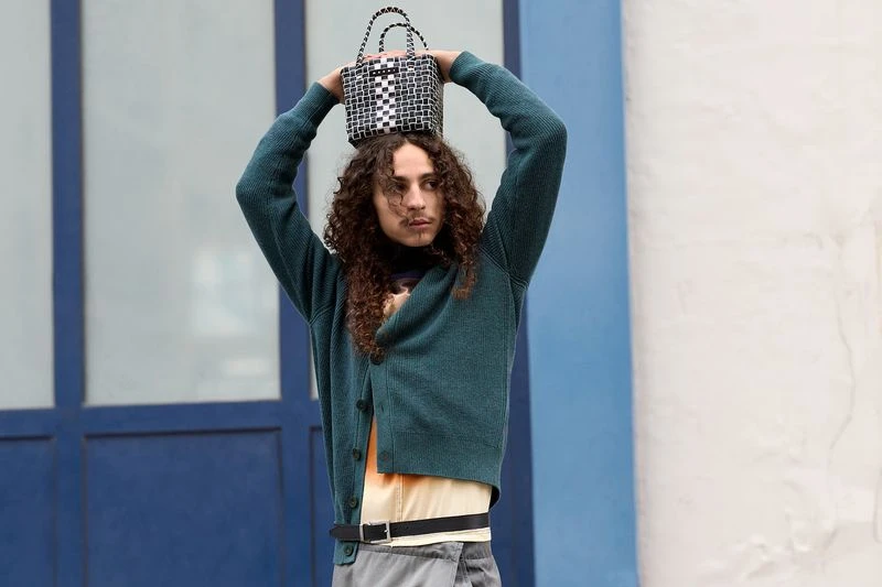 A young adult male with long curly hair balances a black-and-white woven bag on his head. He is wearing a green cardigan sweater.