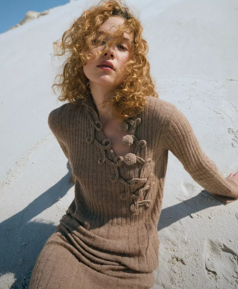 A fashionable woman with curly hair wears a brown knit dress, posing in a sunlit, sandy environment, capturing a relaxed and stylish moment.