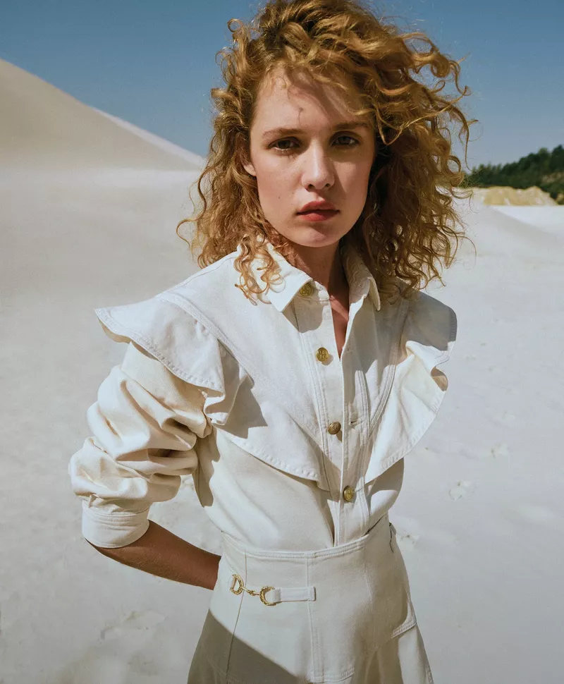 Fashion shot of a curly haired woman in a white denim outfit posing in a bright outdoor desert landscape with sand dunes.