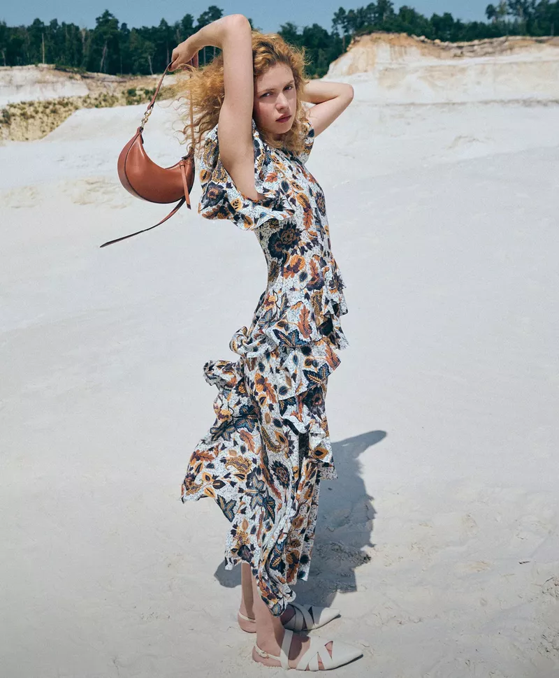 A fair-skinned female fashion model poses in a floral print dress with a brown purse on a neutral white landscape backdrop.