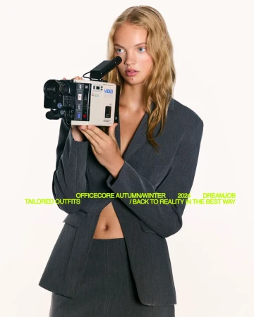 Young woman poses in grey tailored suit with vintage video camera against a white backdrop for fashion editorial, professional headshot, beauty portrait.
