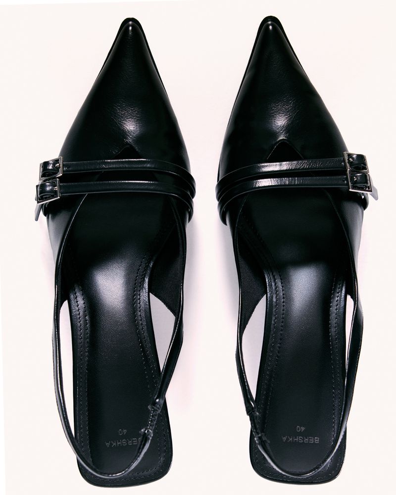 Two black, pointed-toe heeled mules with double-strap detail and silver buckles are displayed on a white surface, showcasing style and design.
