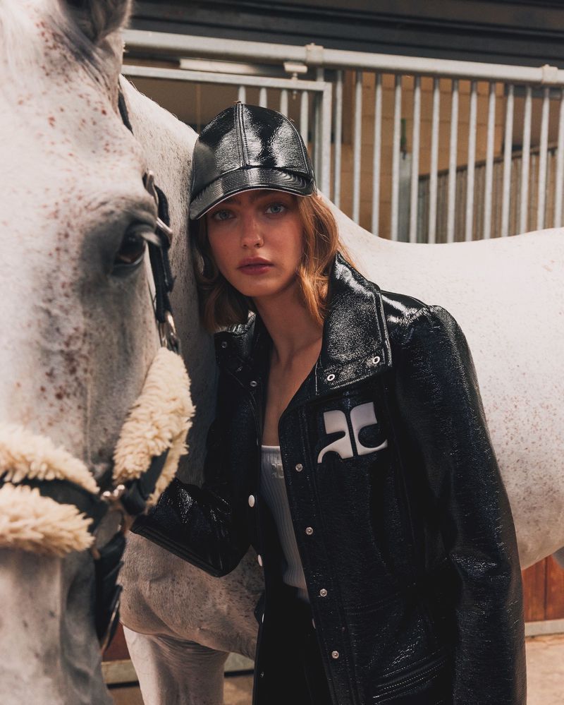 Portrait of a woman in a black jacket and cap standing next to a white horse in a stable with neutral-toned coloring and soft lighting.