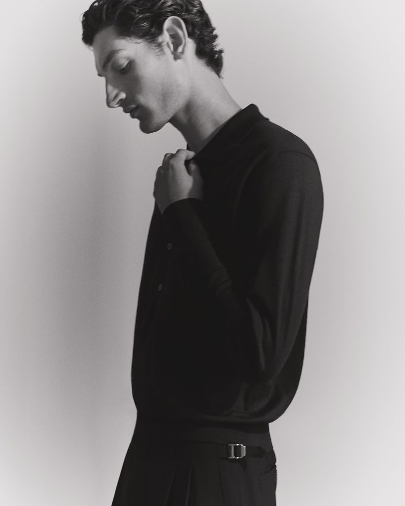 Monochrome portrait of a dark-haired adult man in a long-sleeved polo shirt and pleated pants, in a photo studio with soft lighting.