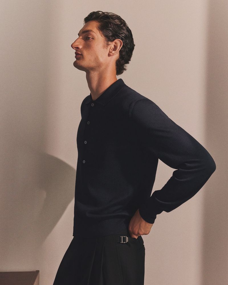 Profile shot of a dark-haired adult male in a dark collared shirt and pleated pants, posing indoors against a neutral wall backdrop.