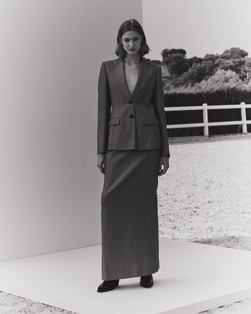 Monochrome portrait of a woman in a pinstripe suit with a long skirt standing in front of a white wall and a rural background.