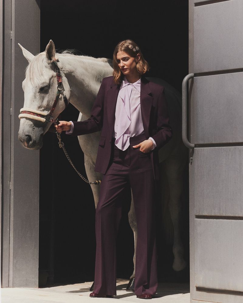 Woman in a plum suit stands beside a white horse in an open doorway, presenting a blend of fashion and equestrian charm.