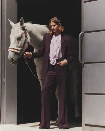 Woman in a plum suit stands beside a white horse in an open doorway, presenting a blend of fashion and equestrian charm.