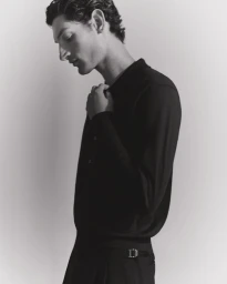 Profile portrait of a male model in a black polo shirt adjusting his collar against a seamless grey studio background.
