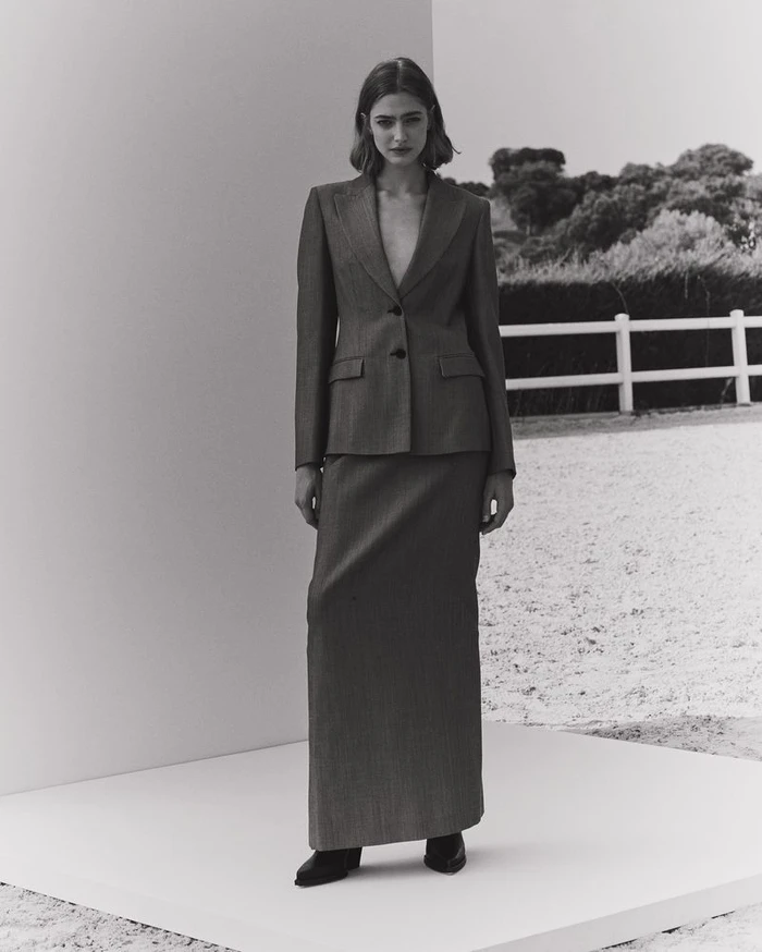 Model wearing a structured blazer and long skirt standing on a white platform outdoors against a landscape backdrop.