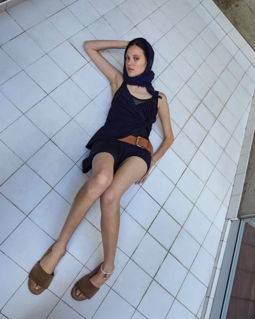 A young woman lies on a white tiled floor wearing a dark blue dress, brown belt, headscarf, and sandals, casually posing for a fashion shoot.