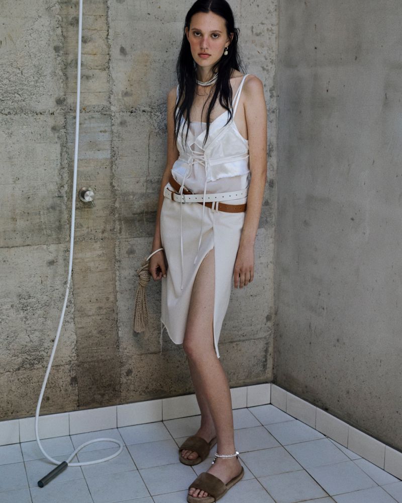 Indoor portrait of a young adult female wearing a white slip dress and brown sandals, posing against a concrete wall and tiled floor.