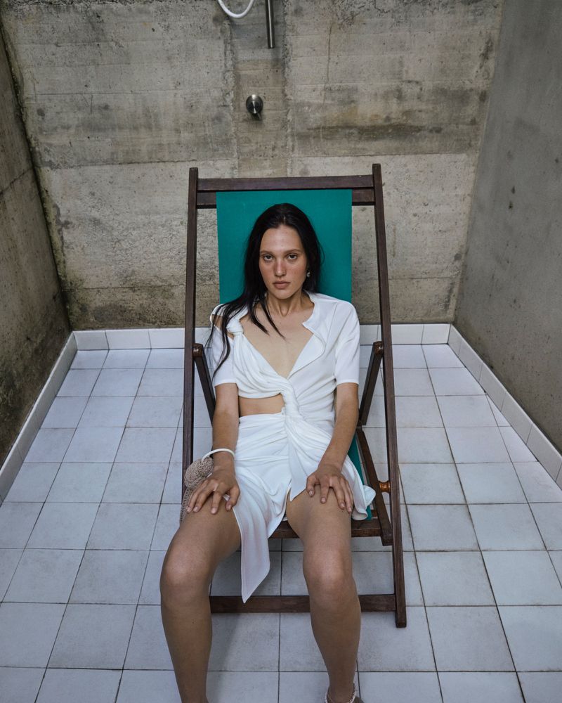 Woman in a white dress sits in a green beach chair in a tiled room with a concrete ceiling and shower head, creating a minimalist fashion photo.