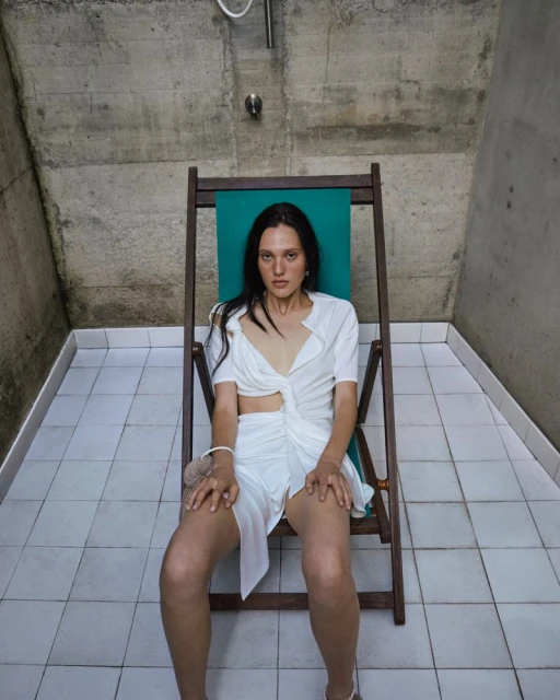 Woman in a white dress sits in a green beach chair in a tiled room with a concrete ceiling and shower head, creating a minimalist fashion photo.