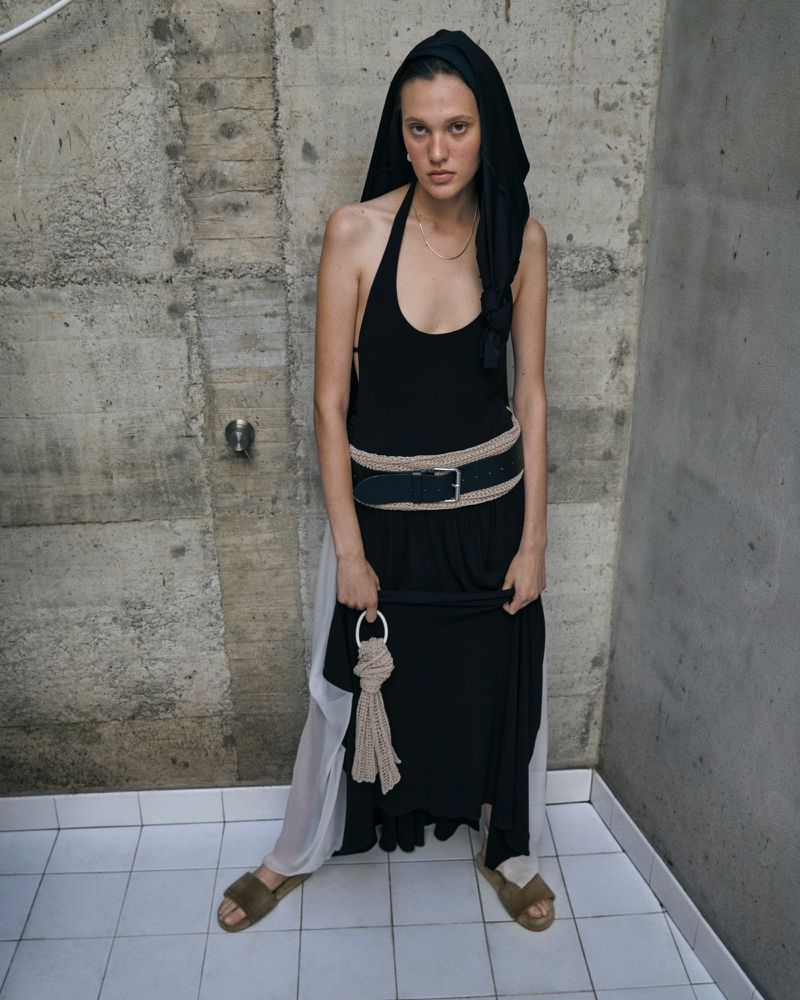 Young woman in black halter dress with hooded scarf and oversized belt poses in front of a textured concrete wall with white tiled floors.