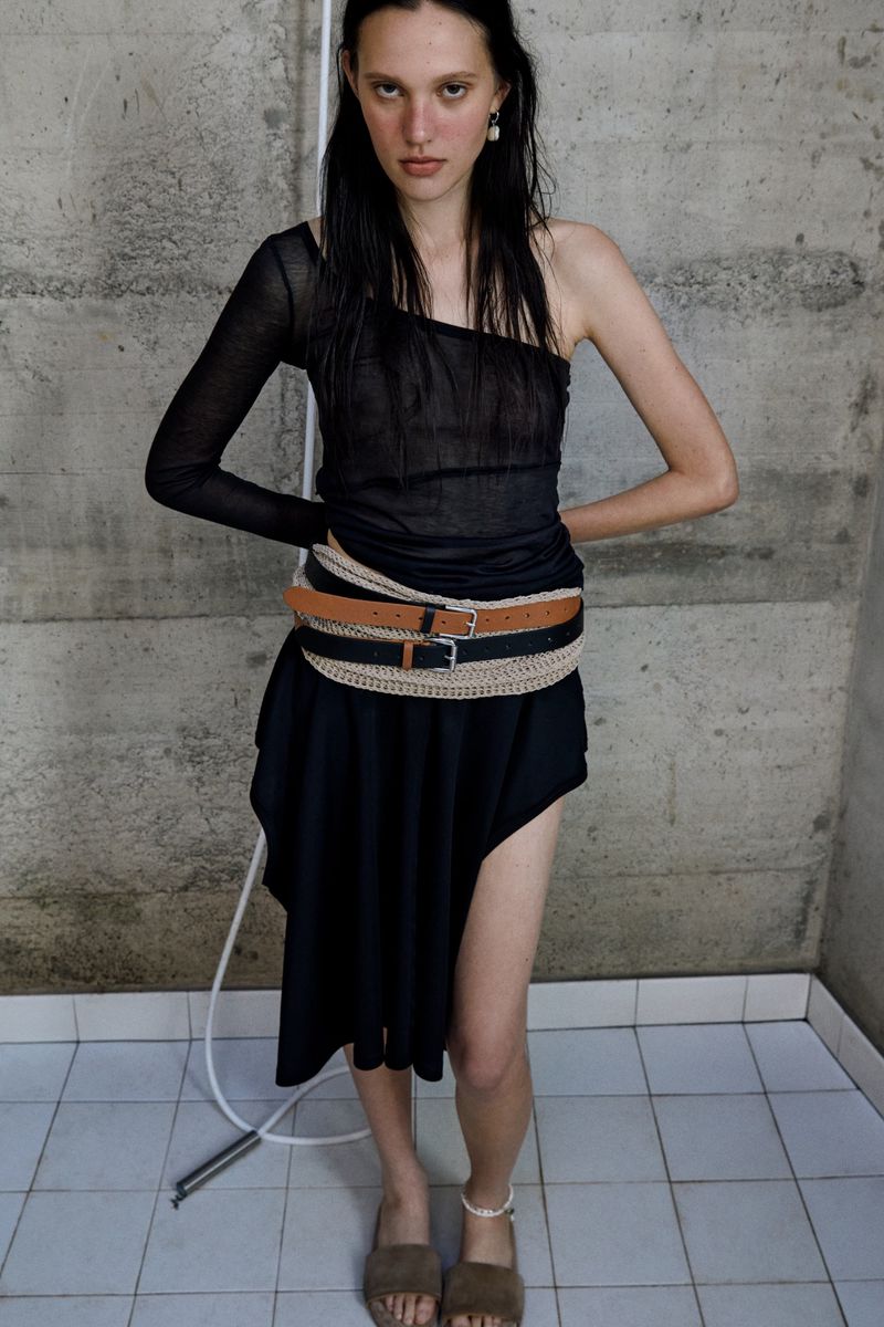 A slender fashion model poses indoors, wearing a one-shoulder top and a skirt with several belts. Tiled floor and concrete wall.
