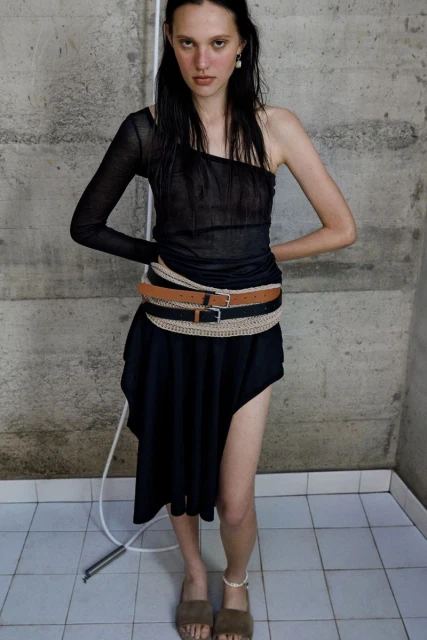 A slender fashion model poses indoors, wearing a one-shoulder top and a skirt with several belts. Tiled floor and concrete wall.