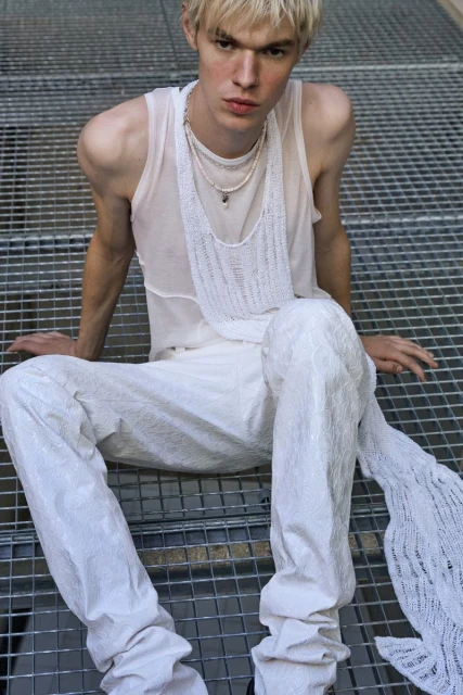 Young adult male model poses while seated on a metal grid surface wearing white top and textured pants, accessorized with layered necklaces.